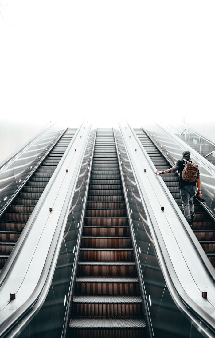 A man with a backpack ascends an indoor escalator under bright lighting.
